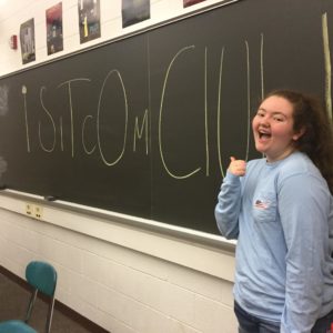 Junior, Maggie Lane, gestures to the Sitcom Club sign on the chalkboard. Lane hopes that by showing Sitcom Club to the students at Community Day, it will help boost students interests in the club. 